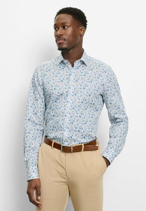 Mann trägt hellblaues langärmliges Hemd mit Blumenmuster, in beigen Hose mit braunem Gürtel gesteckt, steht mit einer Hand in der Tasche.