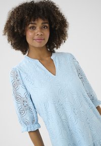 Ljust blå spetsklänning med blommönster, V-ringning och puffiga 3/4-ärmar. Den texturerade tyget har intrikata urklippsdetaljer.