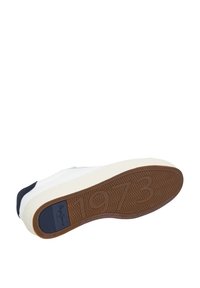 Hvid sneakersål med brunt slidmønster, der viser tallet "1973" og et blåt rektangulært logo-patch.