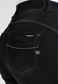 Vaqueros de mezclilla negra con un acabado texturizado, que presentan un bolsillo trasero con costura y un acento de botón de metal. Etiqueta de la marca visible en la cintura.