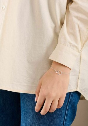 Mână purtând o brățară delicată din argint cu două cercuri interconectate, asortată cu o cămașă crem cu mâneci lungi și blugi albaștri.