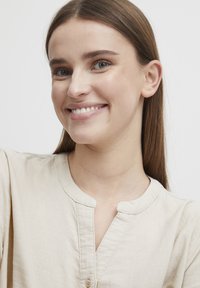 Blouse en lin beige clair avec un col rond, des boutons décoratifs et une texture subtile, portée par une personne souriante sur fond neutre.