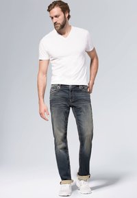 Męskie niebieskie dżinsy z lekkim wyblaknięciem, beżowe przeszycia, prosty krój i podwinięte nogawki, w połączeniu z białą koszulką V-neck i białymi trampkami.