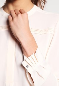Blusa color crema chiaro con sottili pieghe al colletto e ai polsini. Tessuto morbido con una superficie liscia e un dettaglio a scatto argento sui polsini.