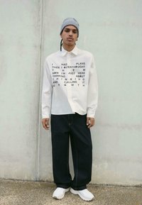Camicia bianca a maniche lunghe con motivo di testo nero; pantaloni in denim scuro a vestibilità ampia; berretto grigio; sneaker bianche; in piedi su uno sfondo neutro.