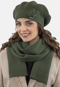 Vihreä neulottu baretti kukkakoristeilla ja siihen sopiva vihreä huivi, jossa on ribat areunat. Molemmissa tuotteissa on sileä rakenne ja mukava ulkonäkö.