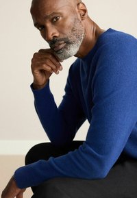 Herrenpullover in Blau aus weichem Strickmaterial, mit Rundhalsausschnitt und langen Ärmeln, vor schwarzem Hintergrund getragen.