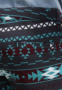 Detail tkaniny s geometrickými vzory v modré, bílé a vínové barvě a malou kapsou na tmavých kalhotách.