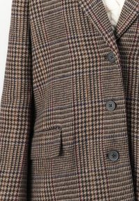 Brun og beige rutete blazer med frontlomme og svarte knapper over en hvit strikkegenser.