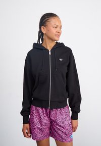 Felpa nera con zip, tasca frontale e cappuccio con cordino, con un piccolo logo a forma di cuore sul petto, abbinata a pantaloni corti con stampa leopardata rosa.