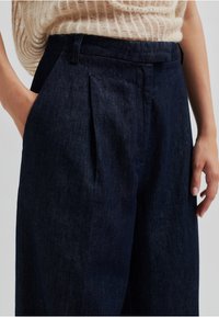 Temno modri jeans hlače s nabranim sprednjim delom, stranskima žepoma in krojem po meri. Mehak tekstur in gladka površina.