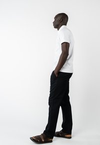 Weißes Kurzarm-T-Shirt, schwarze Hosen, braune Ledersandalen. Das Modell steht seitlich vor einem weißen Hintergrund.