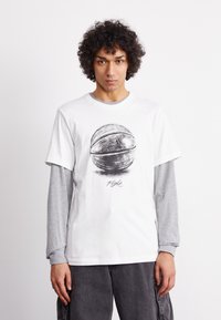 Camiseta blanca de manga corta con un gráfico de baloncesto y el texto "Flight", llevada sobre una camiseta de manga larga gris. Ajuste casual, material de algodón.