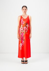 Mujer de pie en un vestido maxi sin mangas rojo con estampado floral en el frente, luciendo sandalias negras de punta abierta sobre un fondo blanco.