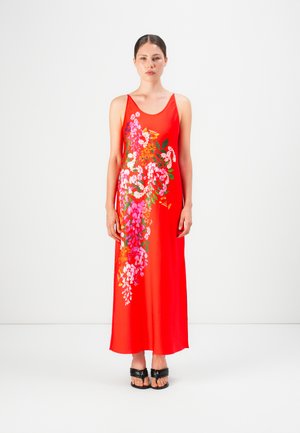 Femme debout dans une robe maxi sans manches rouge avec un motif floral sur le devant, portant des sandales noires à bout ouvert sur un fond blanc.