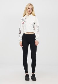 Donna bionda che indossa una felpa bianca corta con schizzi e testo, leggings neri e sneaker nere, in piedi contro uno sfondo bianco.