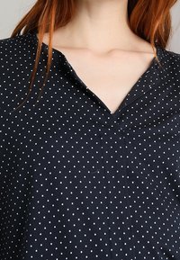 Černá blůza s puntíky, V-výstřihem, krátkými rukávy a lehkým materiálem; malé bílé puntíky rovnoměrně rozložené po povrchu.