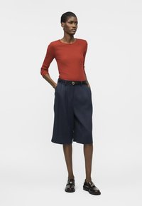 Femme debout, les mains dans les poches, portant un haut à manches longues couleur rouille, un short large bleu marine et des mocassins noirs sur fond blanc.