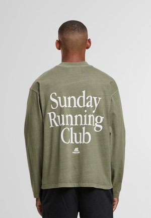 Mand med kort hår iført en olivengrøn langærmet skjorte med teksten "Sunday Running Club" på ryggen, stående foran en ensfarvet baggrund.