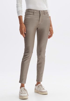 Pantalon taupe coupe ajustée porté avec un haut blanc à manches longues et des baskets blanches, debout devant un fond clair uni.