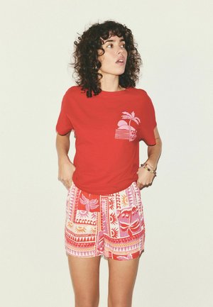 Vrouw met krullend haar draagt een rood T-shirt met een palmboomgrafiek en kleurrijke korte broek met patroon, staand tegen een effen lichte achtergrond.