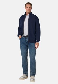 Marinblå stickad zip-up kofta, vit randig skjorta under, blå jeans, ljusa sneakers, ståendes med händerna i fickorna.