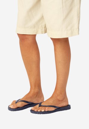 Benen met beige shorts en marineblauwe geweven teenslippers met smalle bandjes, staand op een witte achtergrond.