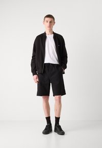 Schwarze Bomberjacke über einem weißen T-Shirt, kombiniert mit schwarzen strukturierten Shorts und schwarzen Turnschuhen. Rotschattierte Socken sind sichtbar.