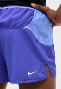 Εγγύς λήψη ατόμου που φοράει μωβ σορτσάκια Nike με λεπτομέρεια φωτεινού μπλε τσέπης και χέρι ακουμπισμένο στο ισχίο του.