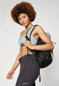 Mochila leve com tecido cinza e detalhes pretos, usada nas costas de uma pessoa. O indivíduo está vestido com um sutiã desportivo estampado e leggings pretas.