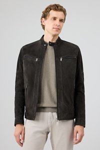 Homme portant une veste en suède noire à fermeture éclair par-dessus un pull côtelé beige et un pantalon de couleur claire, sur un fond gris uni.