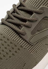 Scarpa sportiva verde scuro con tomaia a trama testurizzata, lacci a contrasto e un'accento in plastica moldata sul lato.