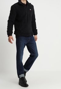 Sudadera de forro polar negra con cuello de media cremallera y superficie texturizada; combinada con pantalones de mezclilla oscuros y botas negras con cordones.