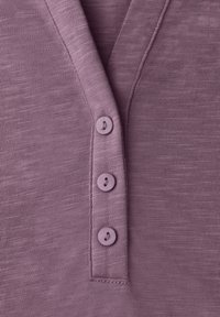 Nahaufnahme einer lila Strickhemdleiste mit drei passenden Knöpfen und sichtbaren Nähten auf strukturiertem Stoff.