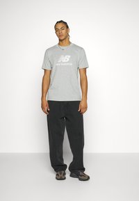 Pilkos medvilnės marškinėliai su "new balance" logotipo, derinami su laisvais juodais kelnėmis ir rudos spalvos raštuotais batais. Paprastas dizainas ir laisvas siluetas.