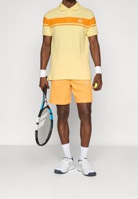 Geel poloshirt met oranje strepen, oranje shorts en witte tennisschoenen. Houdt een tennisracket vast met een blauw en zwart ontwerp.