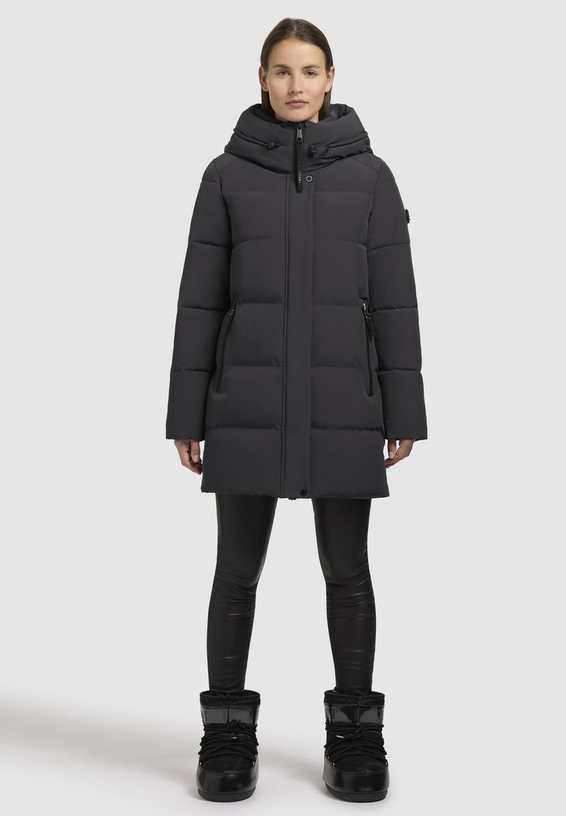 Cappotto invernale imbottito nero con colletto alto, cappuccio, zip nascosta e tasche laterali. Indossato con leggings neri aderenti e stivali neri lucidi.