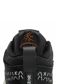 Baskets noires avec matériaux texturés, featuring un accent orange sur la languette du talon. Couture blanche et une étiquette avec le détail "HARDER ONE".