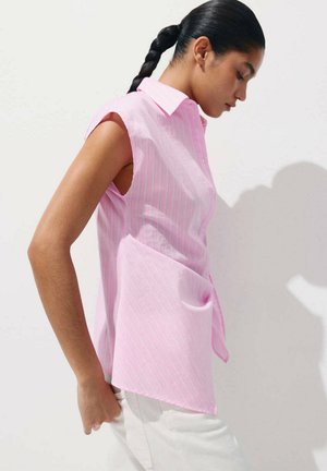 Femme aux cheveux tressés portant une chemise rose rayée sans manches et un pantalon blanc, regardant vers le bas sur un fond clair uni.