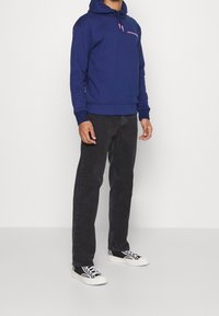 Sudadera azul marino con cordones ajustables, con detalle del logo en el pecho. Combinada con jeans negros y zapatillas en blanco y negro.