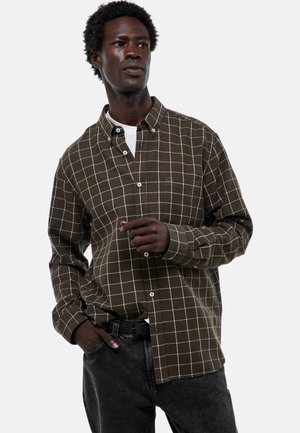 Chemise à carreaux brune avec des lignes blanches, col boutonné, manches longues et poche poitrine, portée avec un jean en denim foncé.