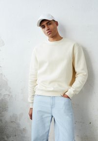 Kremfarget hettegenser med rund hals og ribbestrikkede mansjetter, parret med lys blå jeans. Bakgrunnen har en teksturert hvit vegg.