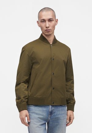 Jeune homme portant une veste bomber vert olive avec col et poignets côtelés, associée à un jean bleu clair, debout devant un fond uni.
