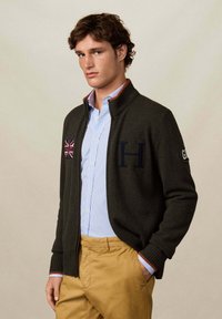 Tmavě zelený svetr na zip s ribbanými manžetami, se velkým nášivkou "H" a union jack na hrudi, nošený přes světle modrou pruhovanou košili.