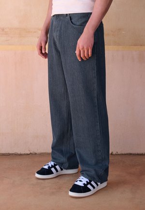 Persona che indossa jeans scuri a gamba larga e sneakers nere con strisce bianche, in piedi su un pavimento beige davanti a una parete beige.