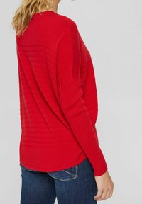 Kvinde iført en rød struktureret langærmet sweater og blå jeans, vist fra ryggen mod en ensfarvet lys baggrund.