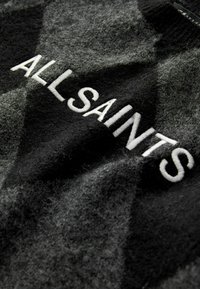 Primo piano di un tessuto di lana con un motivo a scacchi nero e grigio e ricamo bianco che dice "ALL SAINTS" sulla superficie.