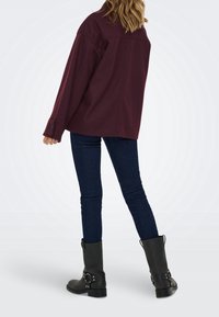 Femme portant un jean skinny bleu foncé, des bottes noires mi-mollet avec des sangles et une veste bordeaux ample, debout de dos.
