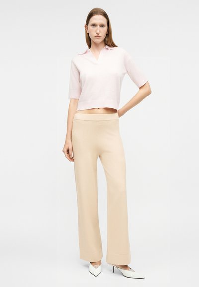 Νεαρή γυναίκα στέκεται με το χέρι στη μέση, φορώντας ανοιχτό ροζ κοντομάνικο πόλο, μπεζ παντελόνι με φαρδιά μπατζάκια και λευκές μυτερές γόβες.