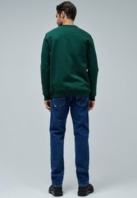 Felpa verde con polsini a coste e collo rotondo, abbinata a jeans blu. I jeans presentano un classico design a cinque tasche e cuciture visibili.